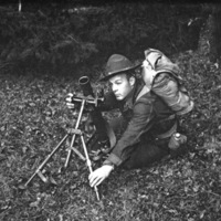 A soldier surrounded by bushes and trees squatting down next to a mortar and adjusting it on its tripod.