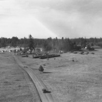 An view from on high and at a distance of a semi-permanent military camp in a wooded clearing.  A dirt road is in the foreground leading into the camp.
