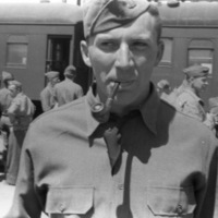 A soldier wearing a garrison cap and smoking a pipe.  Other soldiers and a pair of train cards can be seen in the background.
