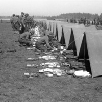 A group of soldiers kneeling or standing and facing a line of tents.  In front of the tents are neatly organized groups of equipment, most likely the contents of the tent and the soldiers' belongings.