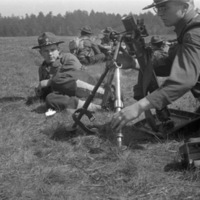 A soldier kneeling next to a mortar and adjusting its tripod.  Another soldier is laying on the grass beside him watching, and another small group of soldiers can be seen nearby in the background.