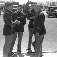Three soldiers listen to a fourth man talking to them.  All of them are in full military dress uniforms. Various military trucks and jeeps are in the background.