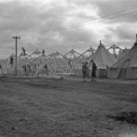 Soldiers working on skeletal wooden structures that make up large tents.  Two completed tents can be seen in the foreground.