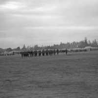 A distant view of a dozens or more soldiers in formation while officers walk in front of them.  A tent camp and milling soldiers can be seen in the background.