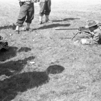 Two soldiers laying down and aiming a large machine gun that is resting on a tripod.  Opposite them is a man laying down facing them and recording them with a camera.  All three are surrounded by other soldiers, who can only be seen from the waist down.