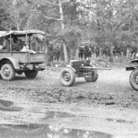 A military truck towing an artillery weapon.  Another truck is following it, and soldiers are seen against a tree line in the background.