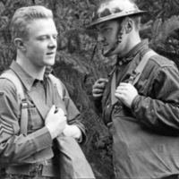 Two soldiers talking with one another.  The one on the left is not wearing their helmet, while the one on the right is wearing theirs.  Trees line the background.