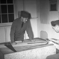 Two soldiers in dress uniform leaning over a carpentry table with various tools on it.