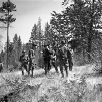 A group of about seven soldiers standing in a lightly wooded area talking with one another.  One of the men can be seen in the background with his back to the camera. 