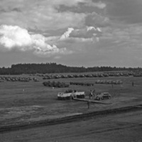 An elevated landscape view of military trucks and soldiers in lines in a wooded clearing. A civilian vehicle is in the foreground, and a stage is set up in the center.