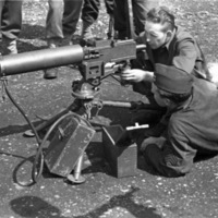 Two soldiers looking and aiming with a large, ground-mounted machine gun.  One is looking from the rear, seeming to aim, while the other lays on the ground watching.