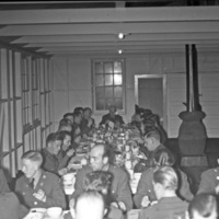 A group of soldiers sitting at a long table having a dinner.   A similar table can be seen partially in the foreground, also occupied by dining soldiers. 