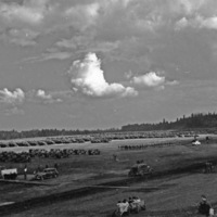 An elevated landscape view of military trucks and soldiers in lines in a wooded clearing.  A few civilian vehicles are in the foreground, and a stage is set up on the left.