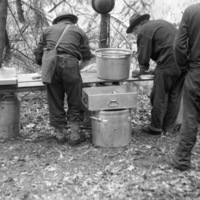 Three soldiers stand with their backs to the camera.  Two of them are leaning over a table that has boxes and pots of food on and beside it.  Trees are seen in the background.