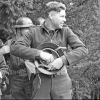 A soldier smiling and looking to the right of the camera while holding and possibly adjusting their helmet.  A few other soldiers are directly behind him with trees in the background.