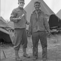 A pair of soldiers standing and looking at the camera humorously.  The man on the left is holding shaving equipment and has shaving cream covering the bottom of his face.  The man on the right appears to be disheveled and dirty, while smiling.
