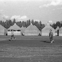 Three soldiers playing basketball in a field.  Tents or small buildings line the background, with trees behind the building.