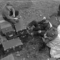 Three soldiers lounging on storage trunks and bedding that is stacked outdoors.