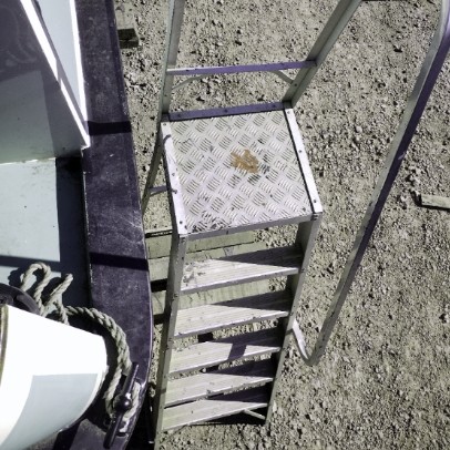 An aluminium (aluminum) stepladder that has 6 steps, standing on a gravel surface and beside the stern of a narrowboat. Several short-cut railroad ties on the bottom of the ladder give it stability.
