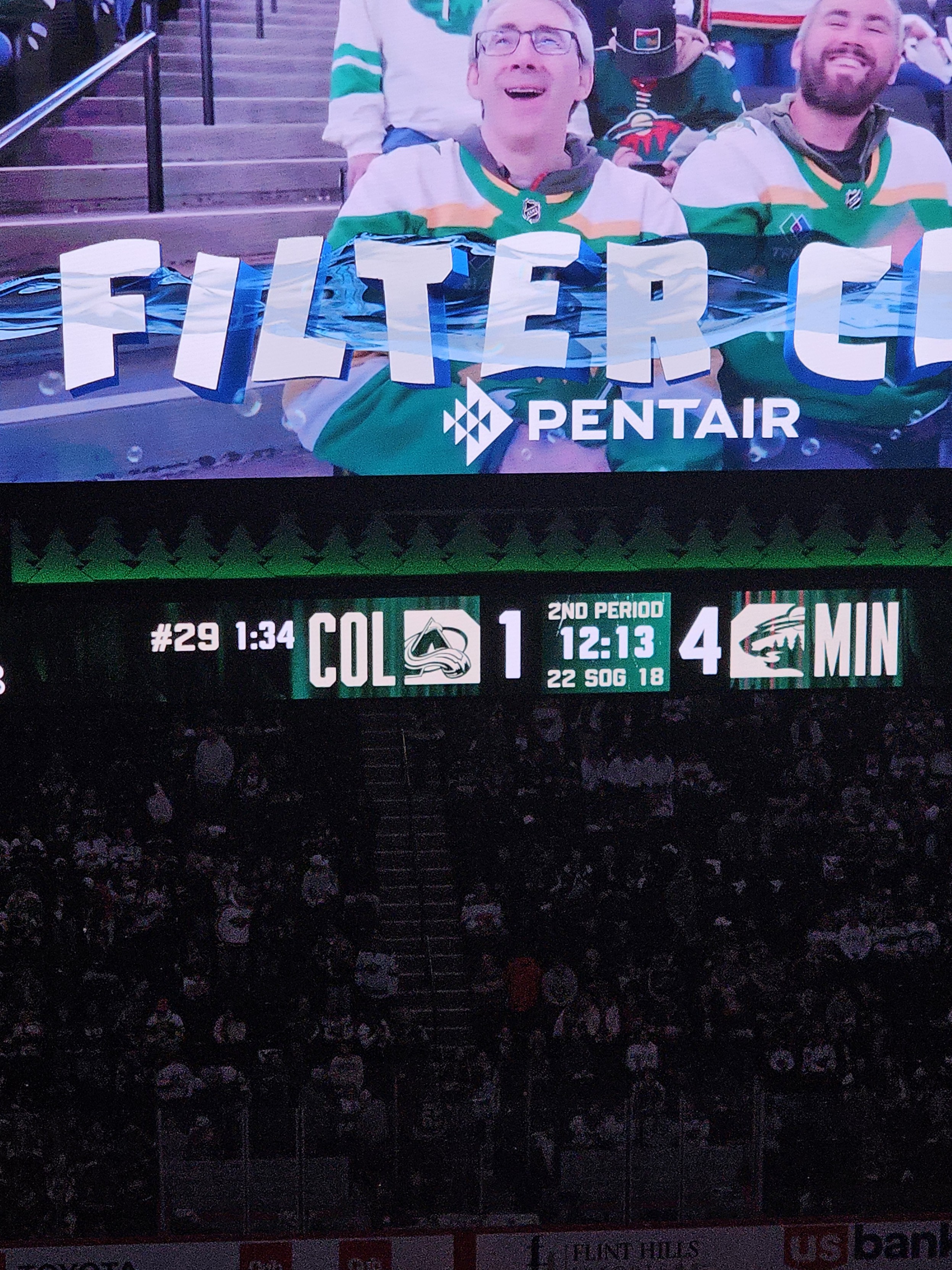A vertical shot of a large jumbotron at a hockey arena during a game. The top half shows two fans on a "Fan Cam" sponsored by Pentair, both wearing green and white jerseys and smiling. Below them, a digital scoreboard displays a score of Colorado (COL) 1 and Minnesota (MIN) 4. The clock shows 12:13 remaining in the 2nd Period. The background is filled with the dark, blurred shapes of a large crowd in the stadium seating.