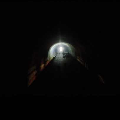This photo was taken 5 minutes from the exit, so 35 minutes through the tunnel.

It shows an arc of light from the boat's headlamp that illuminates part of the tunnel.

There's no footpath in here, just dark water and dark air onside a seemingly endless stone tube build two centuries years ago. Despite the UK drought, there's still some water dripping from above in places.

In the distance is the mouth of the tunnel — a bright blur. The camera didn't capture any sharp detail; in the confined darkness, the small but very bright area ahead was too much for it to focus on. The roof of the narrowboat is also blurry, and fades to dark ess in the photo's foreground, taken from the stern (back of the narrowboat).
