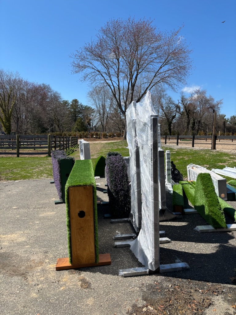 Equestrian show jumping standards and decorative artificial turf fence fillers stored outdoors near a horse arena on a sunny day.