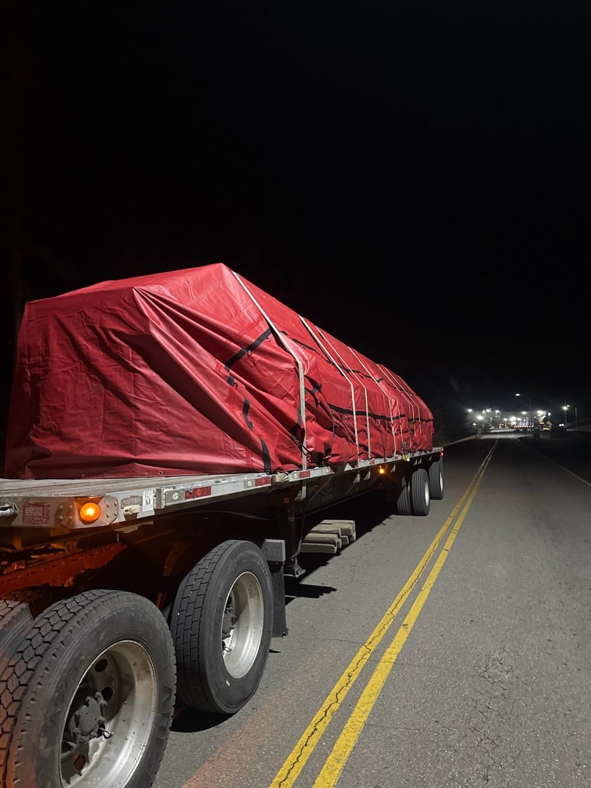 Flatbed semi-truck hauling large red tarp-covered cargo secured with straps on a two-lane highway at night.