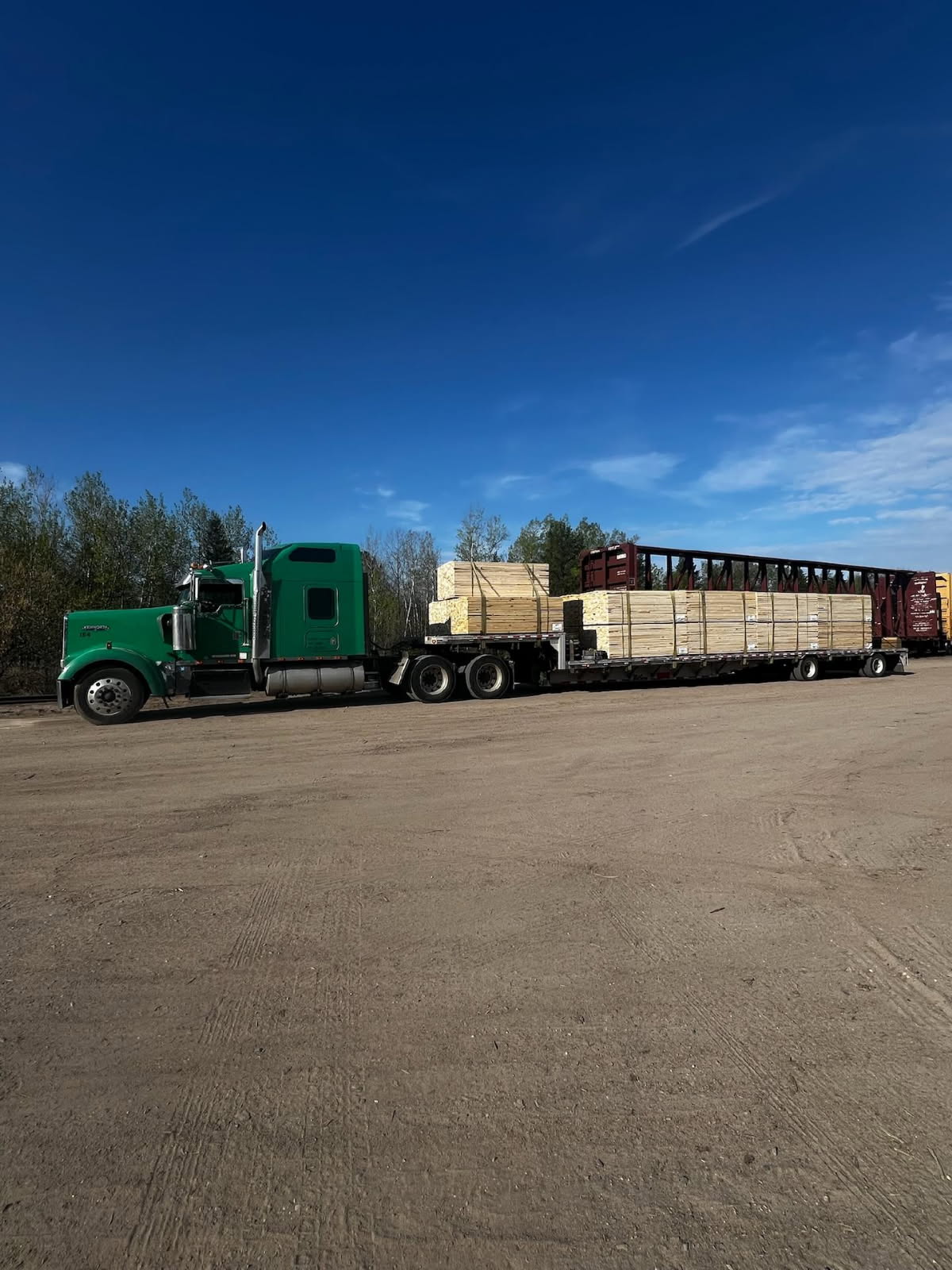 Green semi-truck hauling stacked lumber on flatbed trailer parked on dirt lot beside railway cars under the clear blue sky.