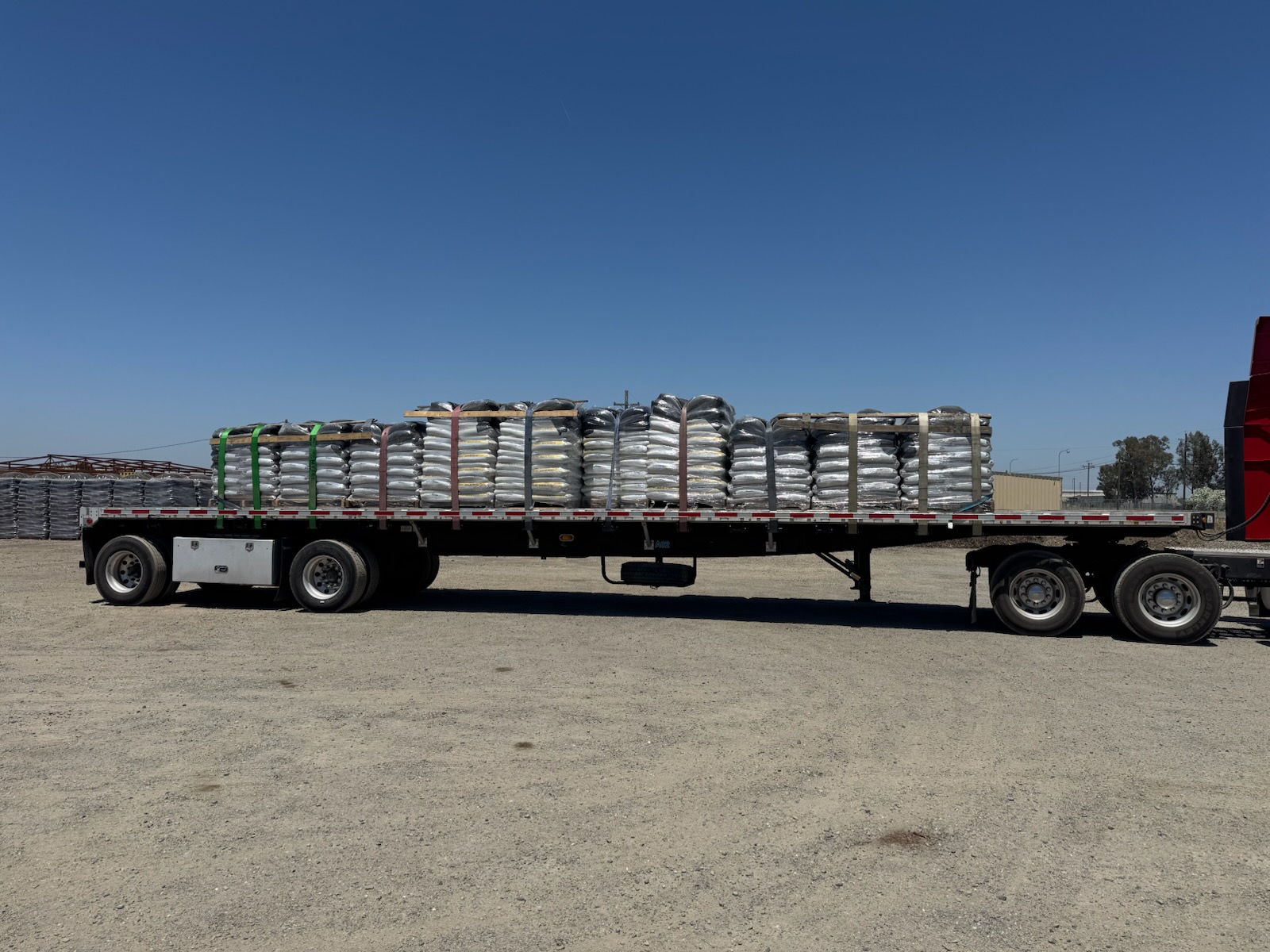 Flatbed semi-truck loaded with pallets of bagged garden materials secured with straps at an industrial yard under clear blue sky.