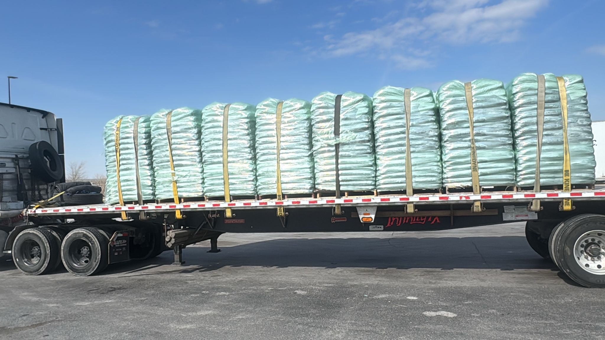 Flatbed semi-truck trailer loaded with green plastic-wrapped bales secured with cargo straps.