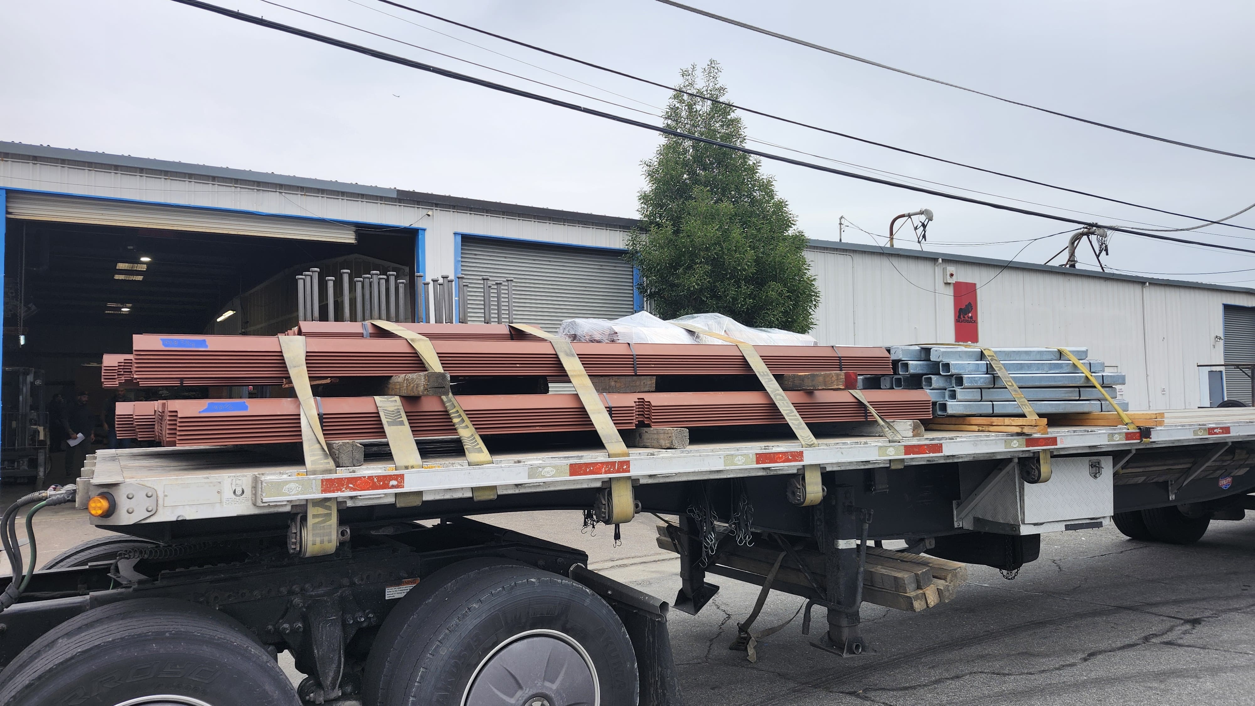 Flatbed truck loaded with steel panels and beams secured with straps outside a warehouse loading dock.