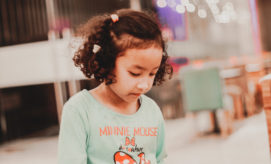 A young girl with curly hair looks down, wearing a turquoise shirt with Minnie Mouse on it. The background is a softly blurred indoor setting with chairs and lights.