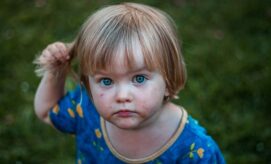A young child with short blond hair holds a strand of hair, wearing a blue shirt with yellow patterns, standing on a grassy lawn, gazing intently.