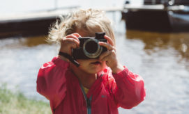 A child in a pink jacket holds a camera, taking a photo near a body of water with boats and a grass-covered shoreline in the background.