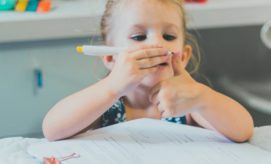 A child holds a pen near her face, giving a thumbs-up over paperwork on a table in a room. She is focused, suggesting involvement with the documents.