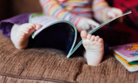 A child with striped clothes sits on a brown couch, flipping through a book using their hands. Another book lies nearby, partially visible. No text is present.