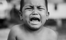 A young child crying with an open mouth, visible tear streaks, and furrowed brows, in an outdoor setting with blurred background elements, suggesting a focus on emotional expression.