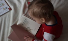 A young child sits on a white surface, wearing a red outfit, looking at an illustrated book. Text in open book: "What are You going to do with your life?" Nearby, a closed book rests.