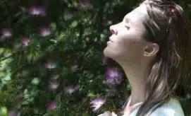 A woman stands peacefully, eyes closed, enjoying sunlight. She is surrounded by greenery with small, purple flowers, creating a serene, natural setting.