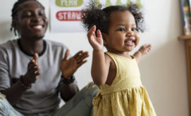 A toddler, dressed in a yellow polka-dot dress, smiles and spreads her arms while an adult in the background claps. Posters are visible on a wall with partial text: "STRAWB...".