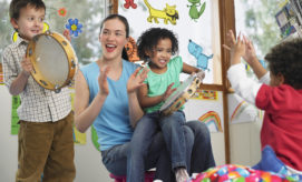 Adults and children play tambourines energetically, clapping and smiling, in a colorful, decorated classroom with animal drawings and rainbows, creating a joyful atmosphere.