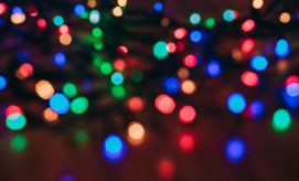 Colorful lights create a soft bokeh effect, displaying a variety of blurred red, green, blue, and yellow dots against a dark background, evoking a festive atmosphere.