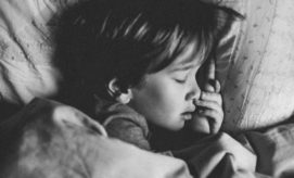 A child sleeps peacefully, with one hand by their face, nestled in cozy bedding. The soft light and textured blanket create a warm, serene atmosphere.