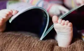 Baby's feet are playfully positioned under an open book on a soft, brown surface, surrounded by blurred, colorful patterns in the background.