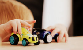 Child's hands push toy cars, one yellow and one blue, across a light wooden surface in a close-up view, with a blurred background.