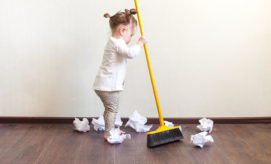 A toddler is sweeping crumpled paper on a wooden floor with a yellow broom, against a plain wall background.