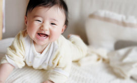 A baby, smiling broadly, is lying on its stomach on a soft bed. The background features pillows and a light, neutral-colored ambiance.