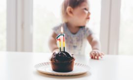 A chocolate muffin sits on a plate with a lit "1" candle, while a baby in patterned pajamas sits behind, near a bright window.