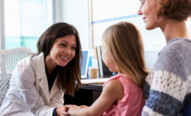 A female doctor in a white coat smiles and holds a young girl's hand while speaking to her in a medical office; a woman sits beside the girl.