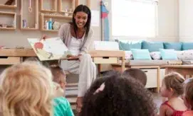 A woman sits on a chair, reading a picture book to a group of young children in a cozy, bright classroom with cushions and shelves in the background.